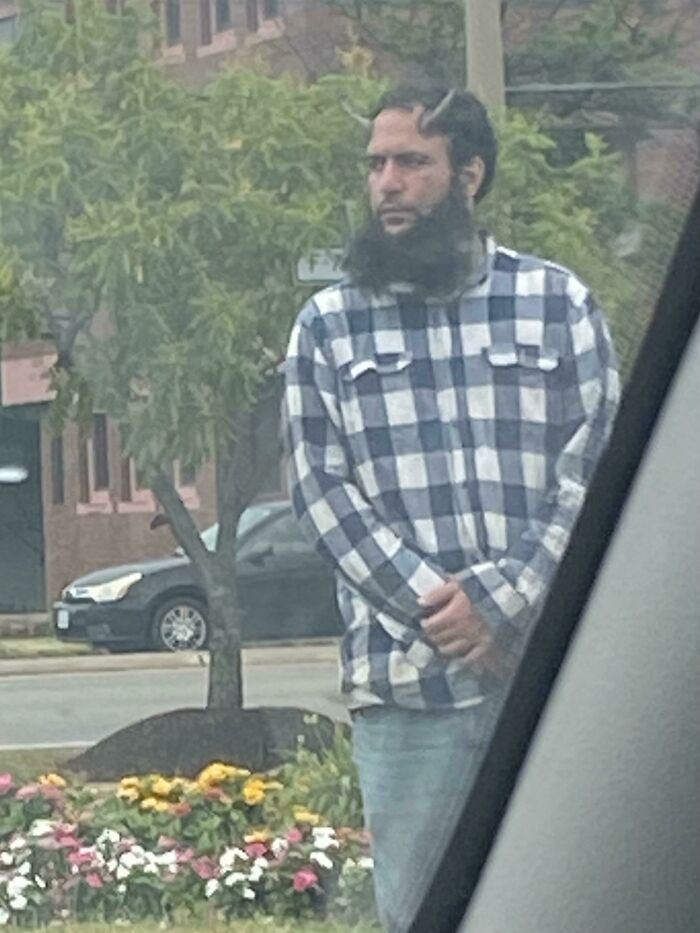 Man in checkered shirt with unusual hair and beard style standing near street with flowers and trees in background.