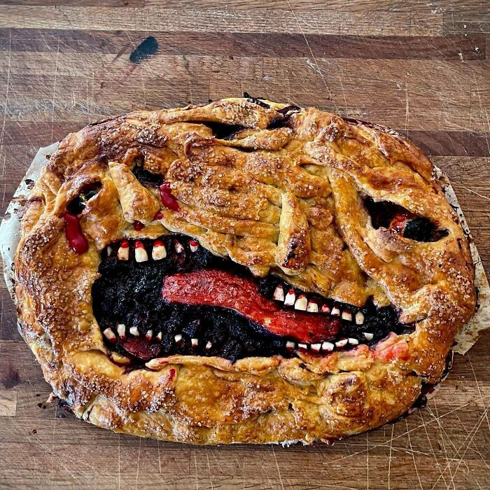 Bizarre pie with a twisted face design, featuring uneven teeth and a red tongue on a wooden surface, showcasing weirdest pics.
