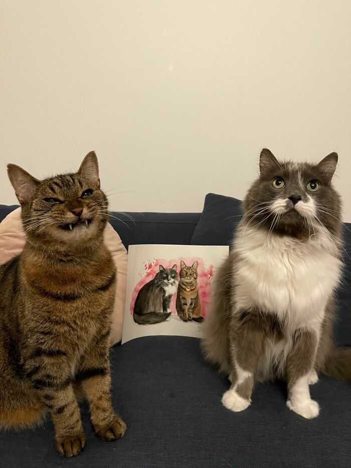 Two cutest cats sitting on a couch with a watercolor painting of cats behind them, charming and heartwarming.