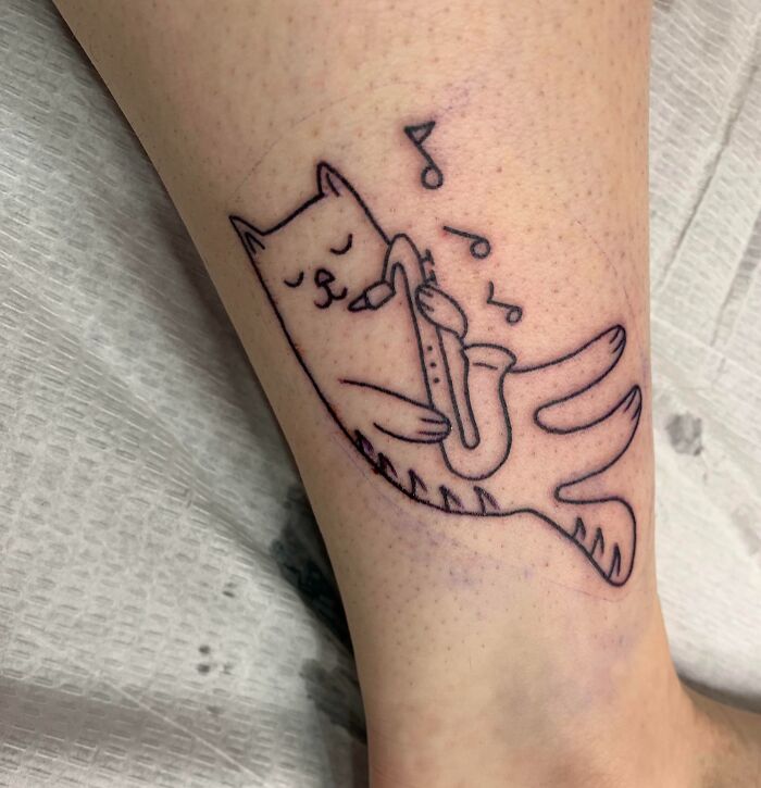 Ankle tattoo of a cat playing saxophone, featuring musical notes.
