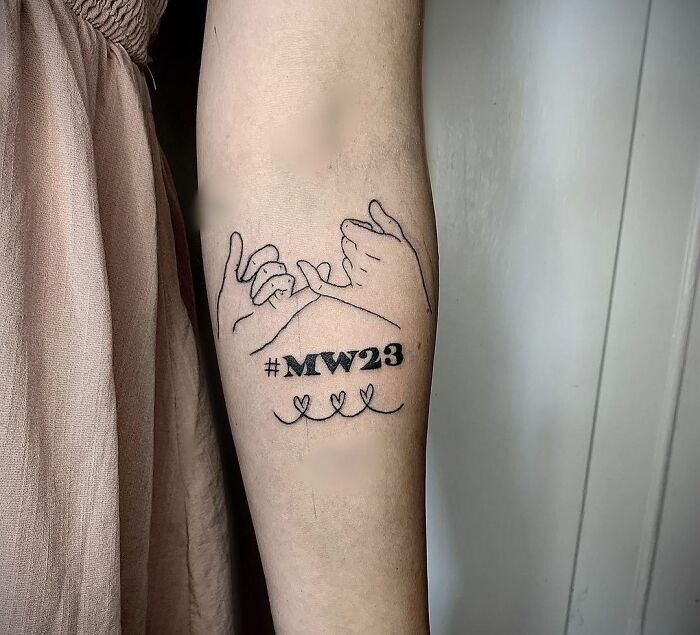 Tattoo of pinky promise with "#MW23" text, symbolizing family honor.