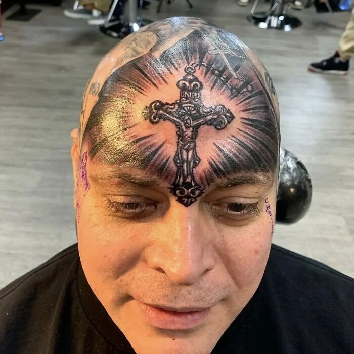 Man with a large cross tattoo inked on his forehead, seated in a tattoo studio.