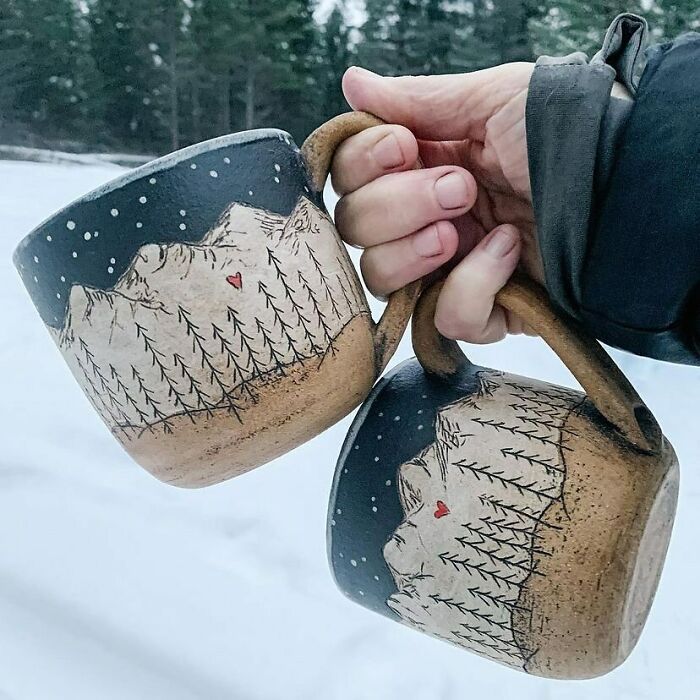 Hand holding two artistic mugs with mountain and tree designs outdoors in a snowy forest setting mug life.