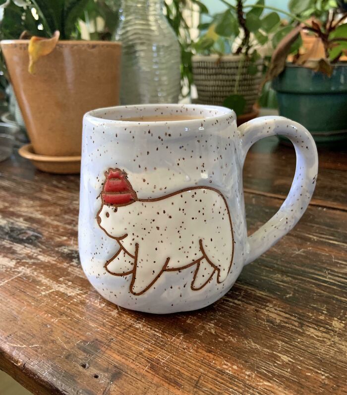 Speckled ceramic mug with a bear wearing a red hat design, placed on a wooden table surrounded by indoor plants.