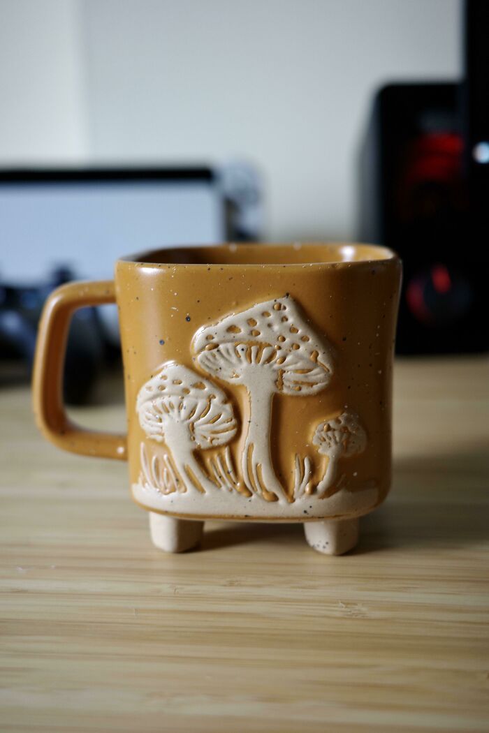 Brown ceramic mug with embossed mushroom design, standing on small legs, placed on a wooden surface in a room.