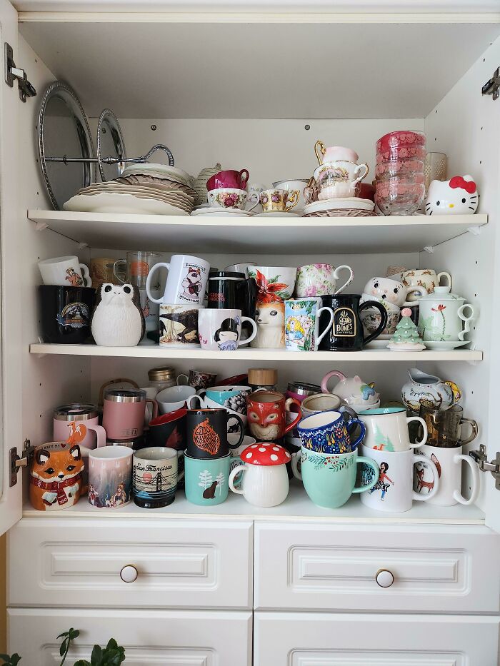 A collection of colorful and uniquely designed mugs displayed on shelves in a white kitchen cabinet.