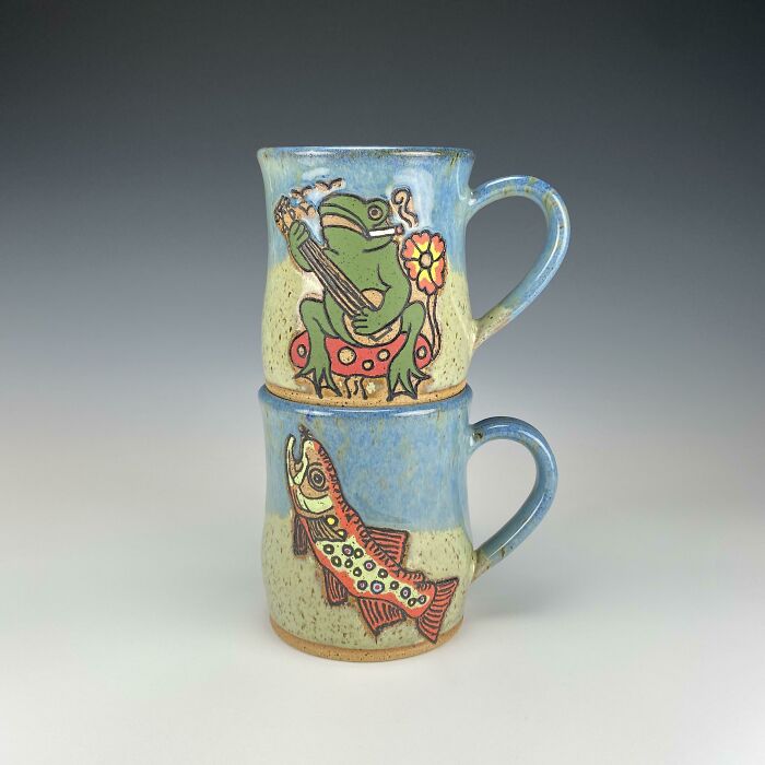 Two ceramic mugs stacked, one with a frog playing guitar, the other with a colorful fish, showcasing mug life designs.