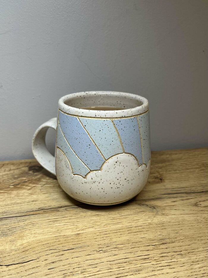 Speckled ceramic mug with blue sky and cloud design sitting on a wooden surface and filled with liquid.