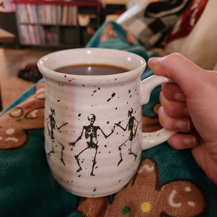 Hand holding a ceramic mug with dancing skeletons design, filled with coffee, showcasing an awesome mug life style.