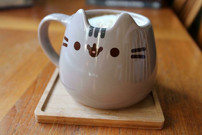 Cat-shaped ceramic mug filled with frothy coffee, placed on a wooden tray on a table, showcasing unique mug design.