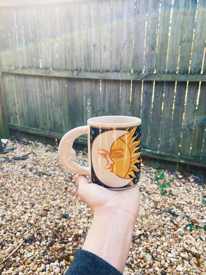 Hand holding a ceramic mug with sun and moon design and a crescent moon shaped handle outdoors on gravel ground.