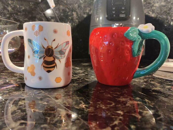 Two colorful mugs on a kitchen counter, one with a bee design and the other shaped like a strawberry, showcasing mug life.