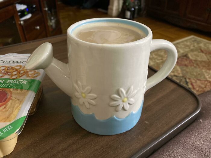 Ceramic watering can mug with daisy decorations filled with coffee on wooden tray beside snack pack.