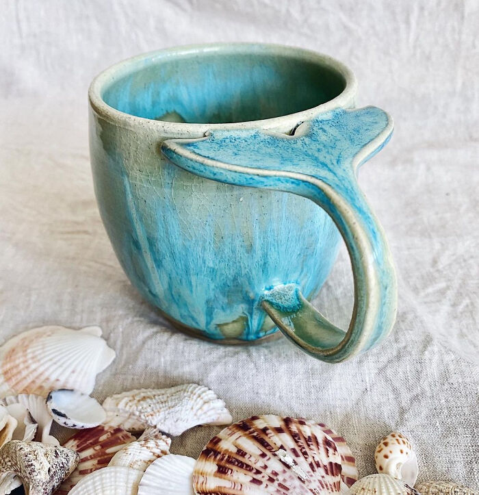 Blue ceramic mug with a whale tail handle, surrounded by seashells, showcasing creative mug life design.