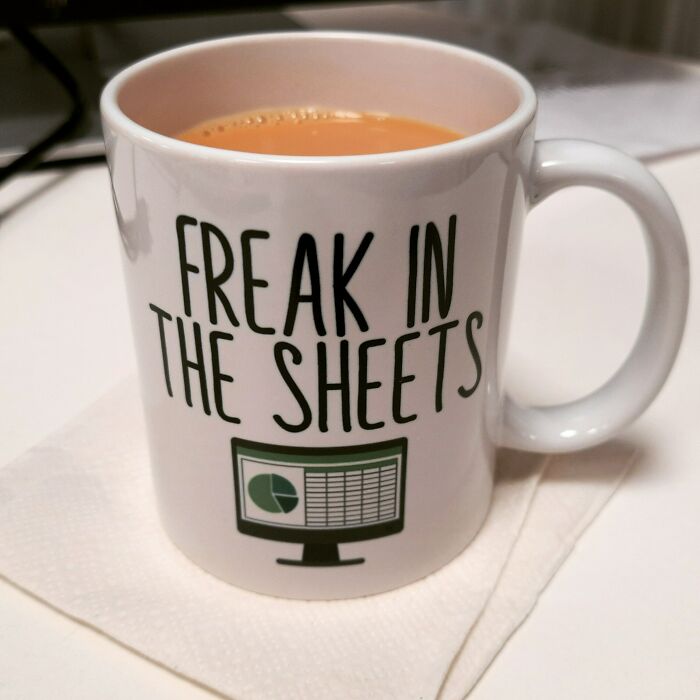 White ceramic mug with humorous text and a spreadsheet graphic, filled with tea on a napkin, showcasing creative mug life design.