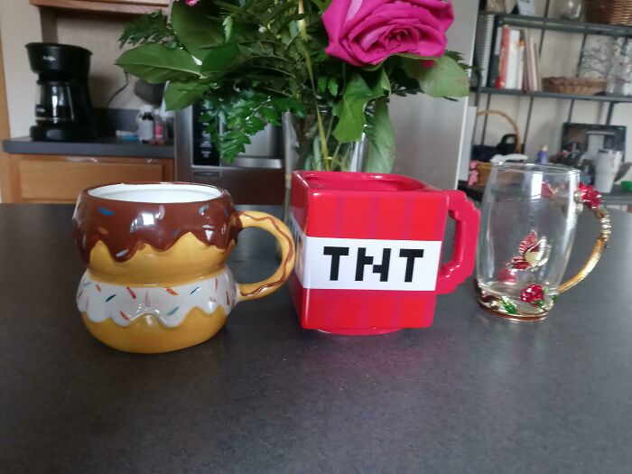 Three unique mugs including a donut, TNT block, and decorative glass with roses on a kitchen counter, showcasing mug life creativity.