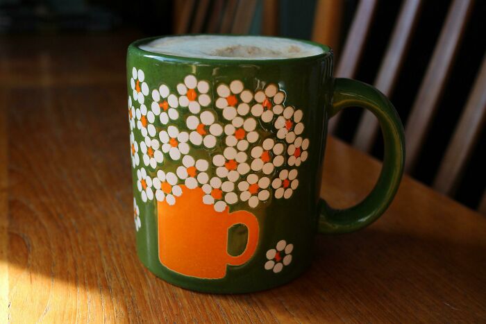 Green ceramic mug with white and orange floral design, filled with frothy coffee, placed on a wooden table.