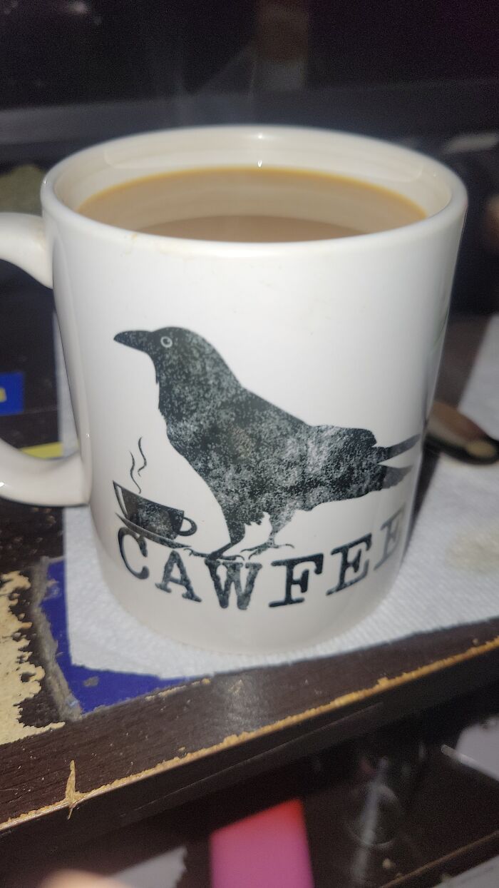 White ceramic mug featuring a black crow and the word cawffee, filled with coffee, on a worn wooden surface.