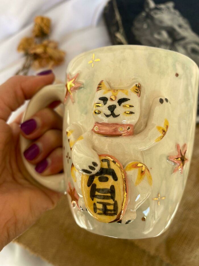 Hand holding a ceramic mug featuring a lucky cat design with raised details and decorative stars, showcasing mug life style.