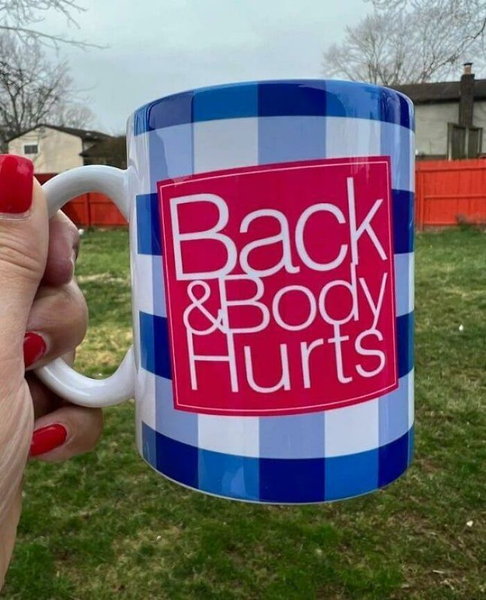 Hand holding a blue checkered mug with pink label reading Back & Body Hurts outdoors in a yard, mug life concept.