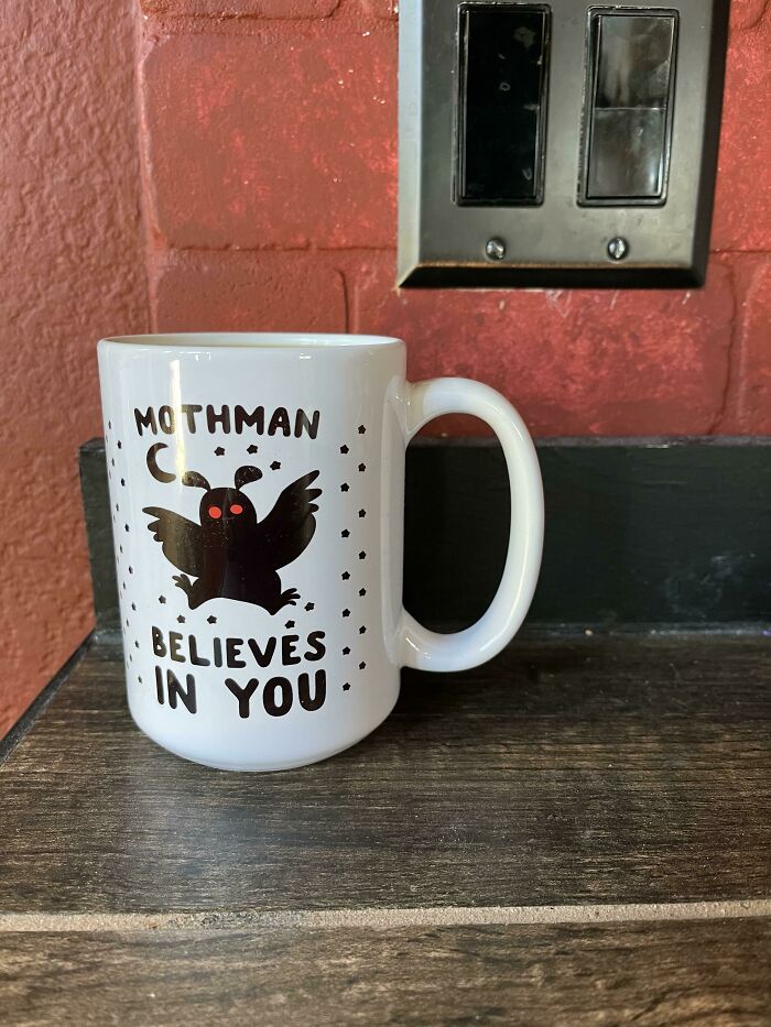 White mug with a Mothman design and red eyes, featuring the phrase believes in you on a wooden surface.