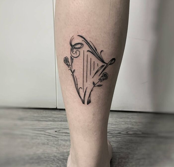 Minimalist music tattoo of a harp with rose accents on a leg.