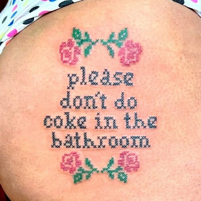 Tattoo with cross-stitch design and text reading "please don't do coke in the bathroom," featuring pink roses.