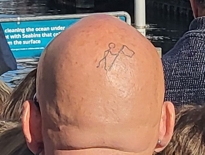 Minimalist tattoo of a stick figure planting a flag on a bald head, illustrating an unusual tattoo choice.