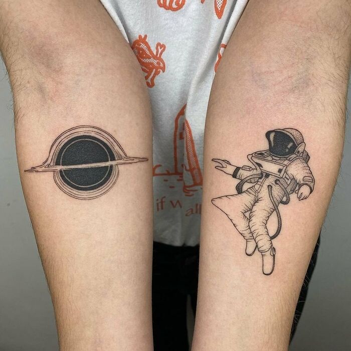 Astronaut and black hole tattoos on forearms, representing science and space exploration themes.