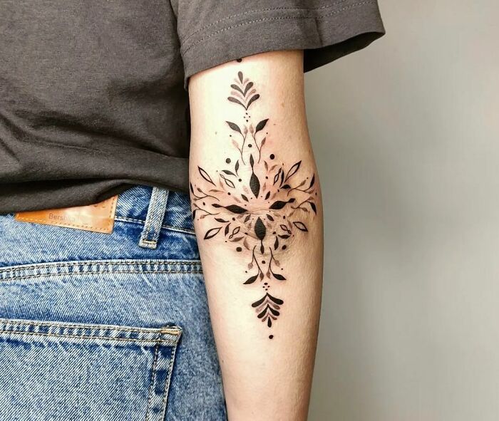Geometric floral elbow tattoo design on a person's inner arm, wearing a gray shirt and jeans.