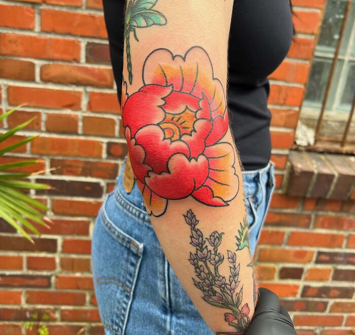 Colorful floral elbow tattoo with red and orange petals, worn by person in black top and jeans, standing by brick wall.