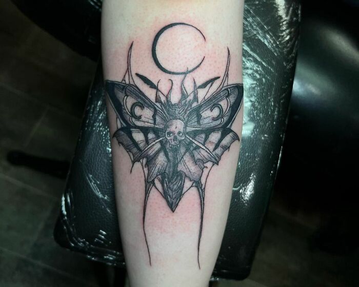 Gothic tattoo of a moth with a skull design and crescent moon on forearm.