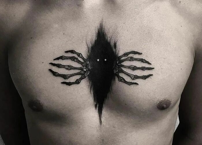 Gothic tattoo of a dark abstract shape with skeletal limbs on a man's chest.