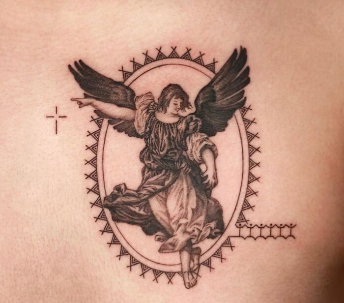 Gothic tattoo of an angel with wings, framed in an ornate oval design with a small cross nearby.