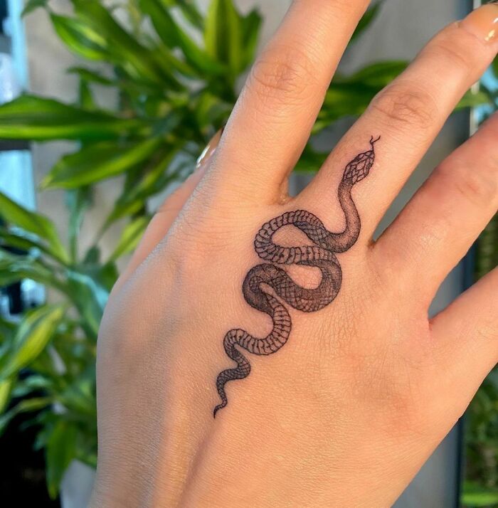 Snake tattoo on a person's hand with green plants in the background.