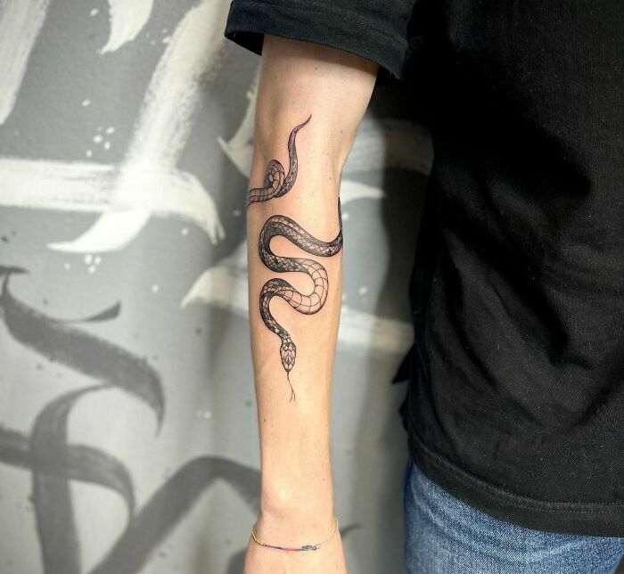 Snake tattoo on person's inner forearm, showcasing detailed black ink design against a blurred background.