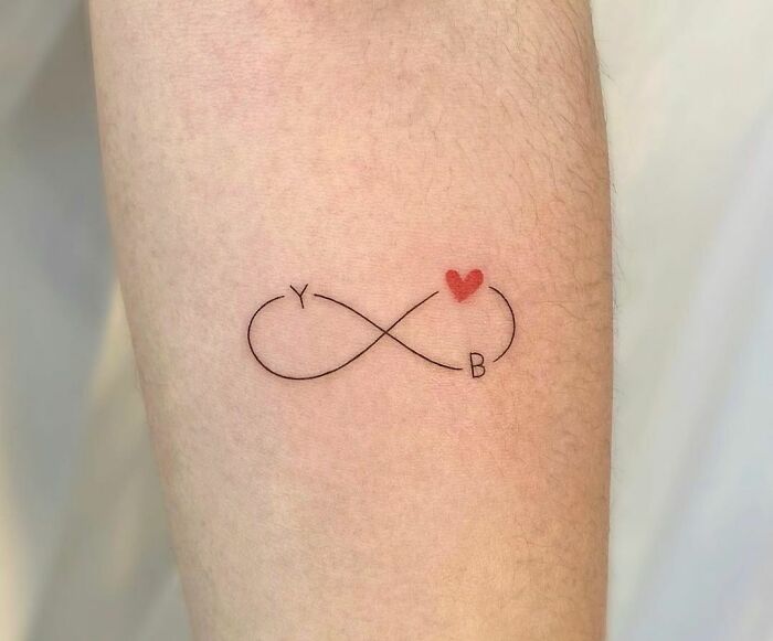 Infinity tattoo with red heart and initials Y and B, symbolizing eternal creativity on inner forearm.