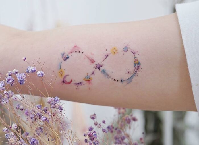Infinity tattoo with celestial and space-themed elements on an arm, surrounded by delicate purple flowers.