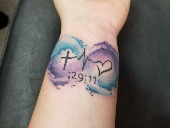 Infinity tattoo with a cross, heartbeat, and heart design on the wrist, surrounded by blue and purple watercolor.