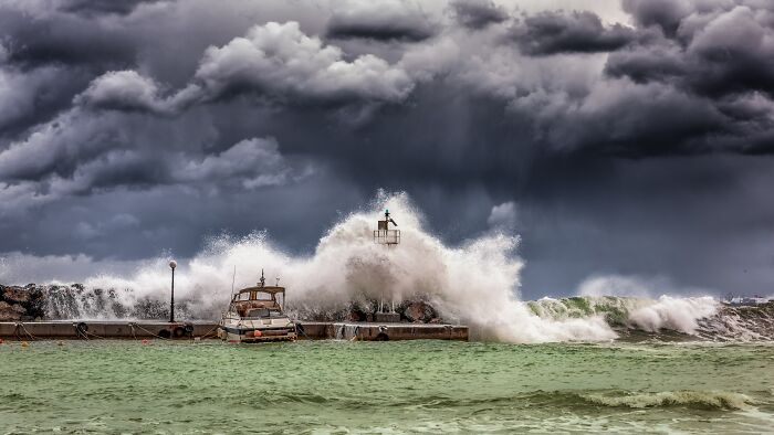 Storm waves crashing over a pier with a boat docked nearby under dark clouds illustrating human stupidity risks.