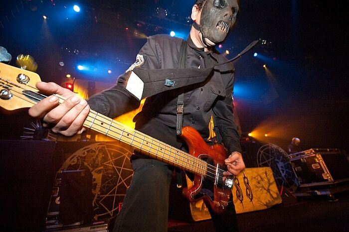 Musician wearing a creepy mask playing bass guitar on stage during a lively rock concert with colorful lighting.