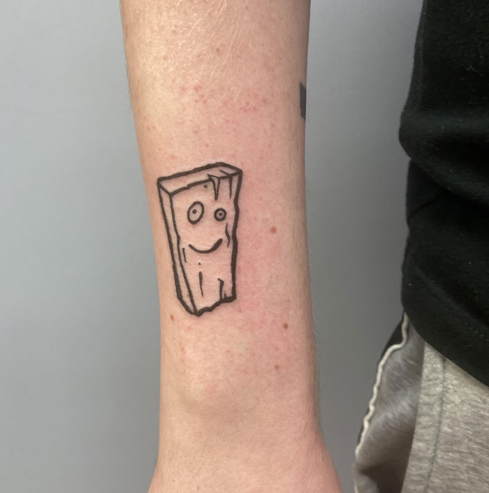 A ‘90s tattoo featuring a cartoon-style smiling plank on an arm.