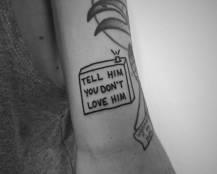 Arm tattoo reads "Tell him you don't love him," offering a powerful mental health message. Black ink design.