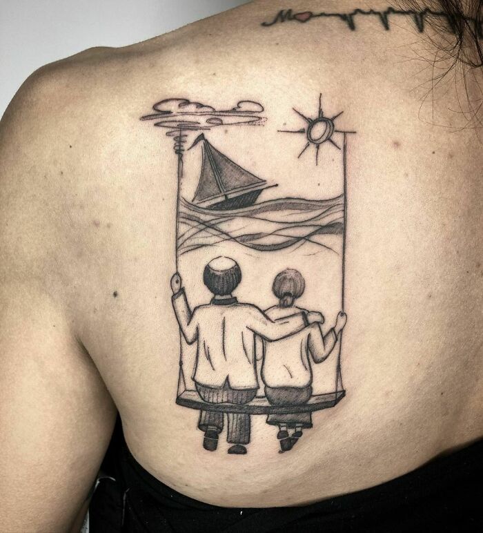 Tattoo of two figures embracing, sitting on a bench with a sailboat and sun, honoring family.