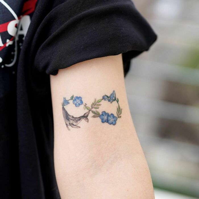 Infinity tattoo with blue flowers and a bird design, symbolizing eternal creativity on an arm.
