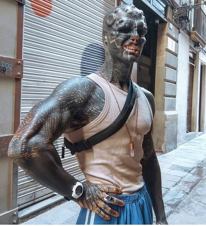 Man with extensive body modifications and tattoos standing in urban setting, wearing a tank top and watch. Man with extensive body modifications and tattoos standing in urban setting, wearing a tank top and watch.