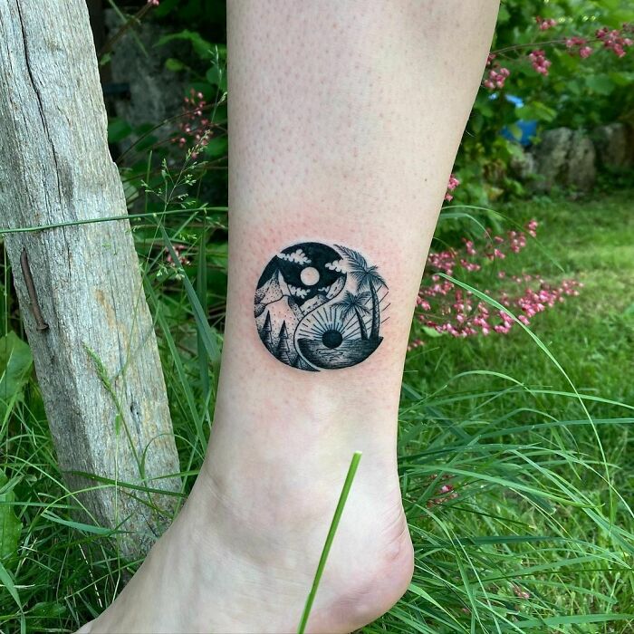 Yin Yang tattoo on leg, depicting a nature scene with mountains and trees, surrounded by grass.