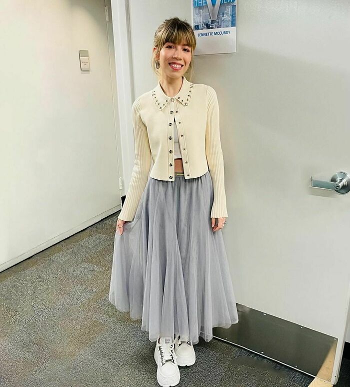 Young woman in a stylish outfit, standing and smiling indoors, related to a personal story about her mother. Young woman in a stylish outfit, standing and smiling indoors, related to a personal story about her mother.