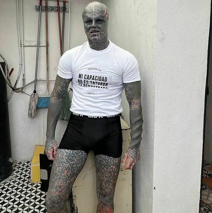 Tattooed individual standing with arms at sides, wearing a white shirt and black shorts, expressing unique self-identity. Tattooed individual standing with arms at sides, wearing a white shirt and black shorts, expressing unique self-identity.