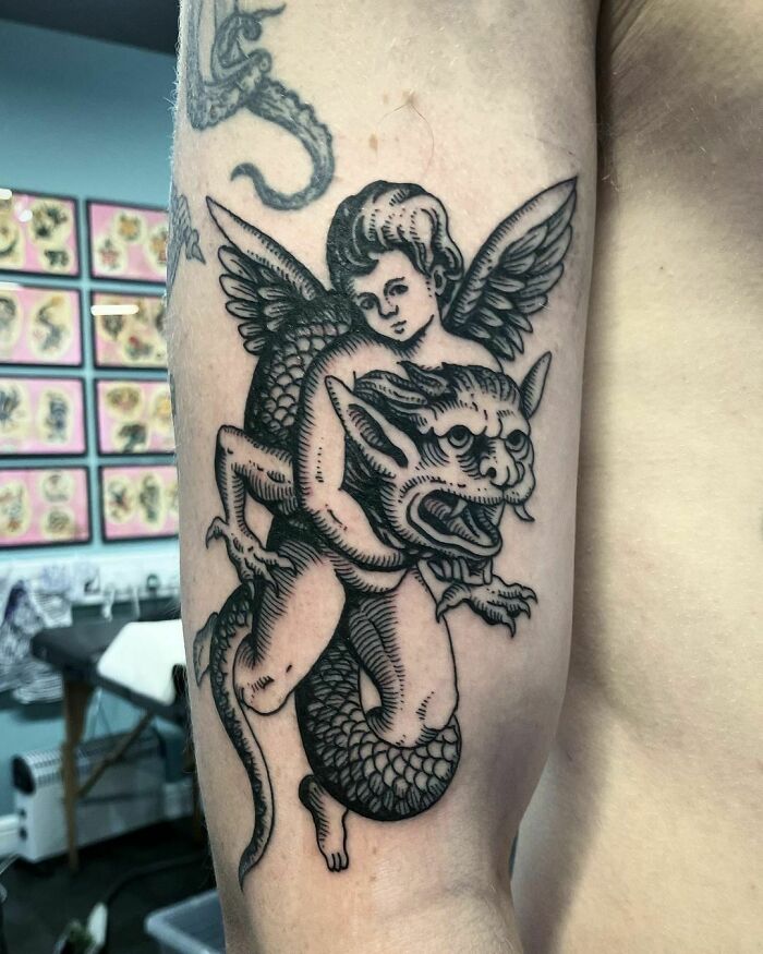Gothic tattoo featuring a winged cherub entangled with a mythological creature on an arm.